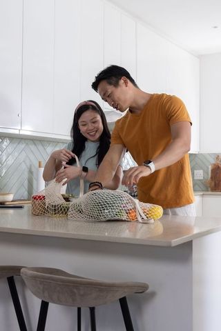 Asian couple unpacking groceries at modern kitchen island with reusable mesh bags