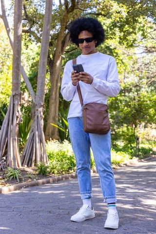 African american man using smartphone on sunlit park path with leather crossbody bag