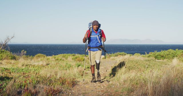 Biracial man with prosthetic leg trekking with backpack and walking poles on coastal path. Long distance walking, fitness, challenge, disability, nature and healthy outdoor lifestyle.