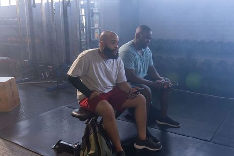 Two african american men sitting on workout benches checking smartphones inside modern gym