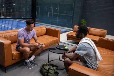 Diverse male friends relaxing on leather sofas before padel match checking phone and planning play