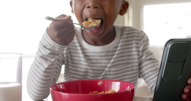 A young boy in a striped shirt is eating breakfast cereal from a red bowl while engrossed in a tablet. A glass of milk is also present on the table. This photo can be used in advertising related to children's nutrition, family routines, or digital devices used at home.