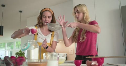 Two friends baking together sifting flour with sieve at sunlit modern kitchen island countertop