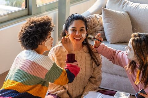 Diverse friends applying facial masks at cozy home spa party, laughing, relaxing together