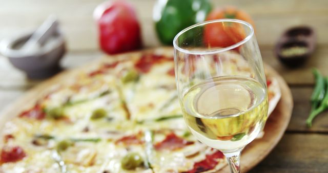 Close-up glass of white wine with Italian pizza arranged on wooden table. Pizza topped with olives, cheese, and bell pepper slices. Bell peppers and tomato blurred in the background for bokeh effect. Ideal for use in food blogs, restaurant menus, gourmet dining promotions, and lifestyle articles.