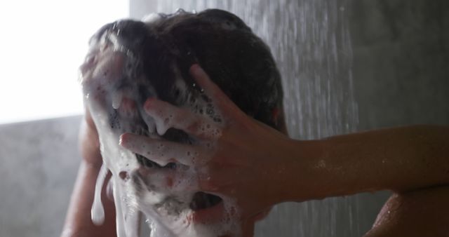 Person washing hair with shampoo, hands creating foam and suds in the shower. Useful for personal care, hygiene, haircare products advertisements, self-care, daily routine visuals.