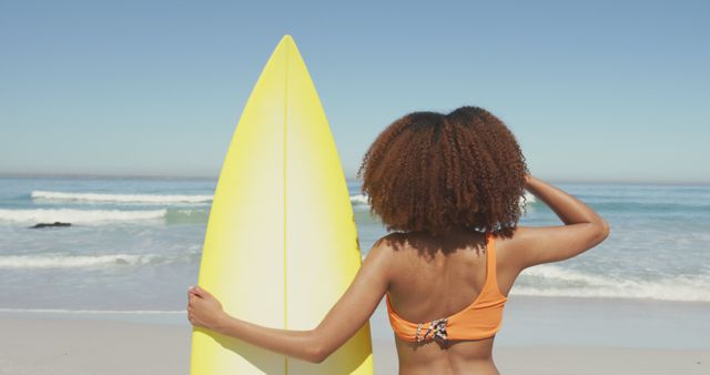 Back view biracial woman standing on beach, holding surfboard and looking at sea. Summer, vacation, relaxation, happy time.