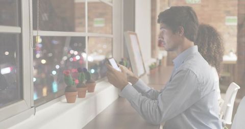 Tapping smartphone at window desk in nighttime coworking space with cacti and monitors