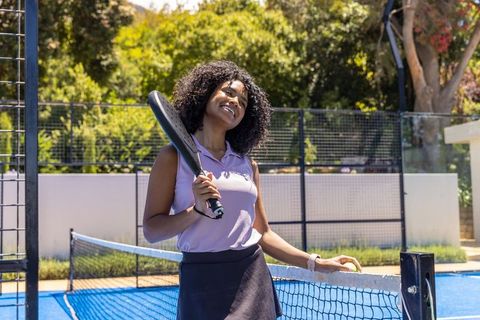 Smiling african woman holding padel paddle and ball on sunny blue court