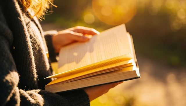 Sunlit woman reading paperback with yellow bookmark at golden hour in cozy knit sweater