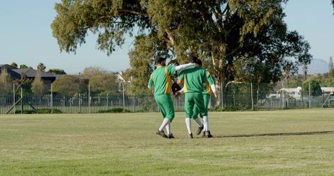 Diverse male baseball teammates walking across grass field embracing after practice