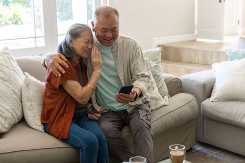 Senior couple sharing smartphone on cozy living room sofa smiling and embracing