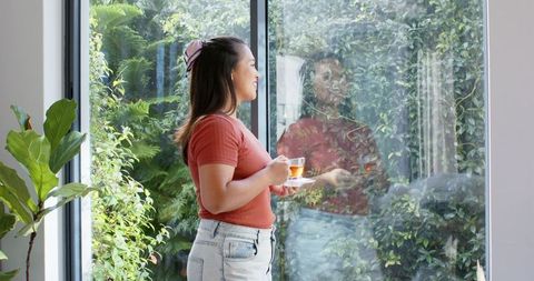 Asian woman holding teacup looking out glass door at lush garden