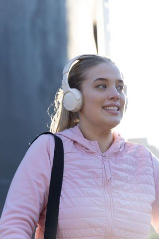 Young woman listening to music on wireless headphones walking in city wearing pink jacket
