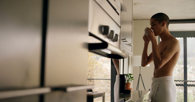 Young person in a towel sipping coffee in a contemporary, bright kitchen. Great for concepts of morning routines, relaxed lifestyle, residential living, self-care, and casual home environments.