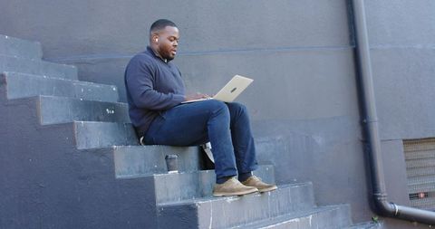 African American man working on laptop on urban concrete steps with coffee and earbuds