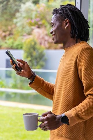 African american man smiling while holding smartphone and coffee mug on patio by pool