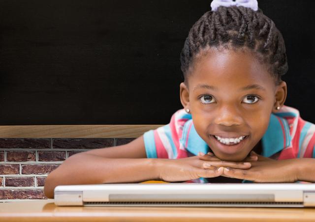 Young African American girl with braided hair smiling and resting her face on a laptop in a classroom. Ideal for educational content, technology in education, school promotions, and child learning materials.
