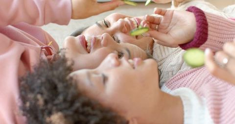 Diverse women lying together sharing cucumber facial slices in cozy pastel self-care moment