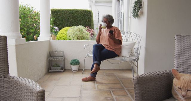 Senior woman relaxes on cozy porch bench with a hot drink and newspaper. Beautifully tended garden and tranquil atmosphere enhance the rustic charm. Ideal for illustrating leisure time, senior lifestyle, outdoor relaxation, peaceful home settings, or promoting health and wellbeing in a casual atmosphere.