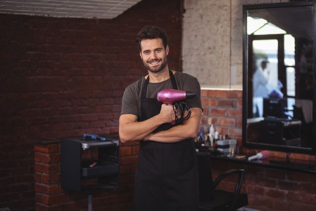 Male hairdresser standing in a modern salon with a brick wall background, holding a hairdryer and smiling confidently. Ideal for use in advertisements for hair salons, grooming products, hairstyling services, and professional training courses.