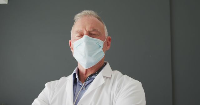 A senior doctor is confidently standing with his arms crossed while wearing a protective mask in a medical clinic. This stock photo could be used for healthcare advertisements, medical articles, health insurance promotions, or hospital brochures, highlighting the importance of professionalism and safety in healthcare environments.
