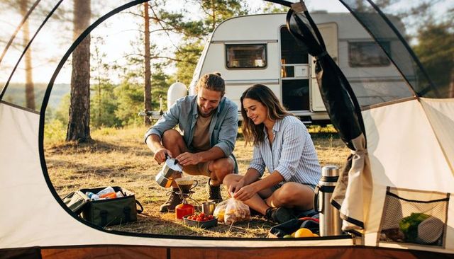 Couple making coffee at campsite beside caravan during golden hour in pine forest, relaxed morning r