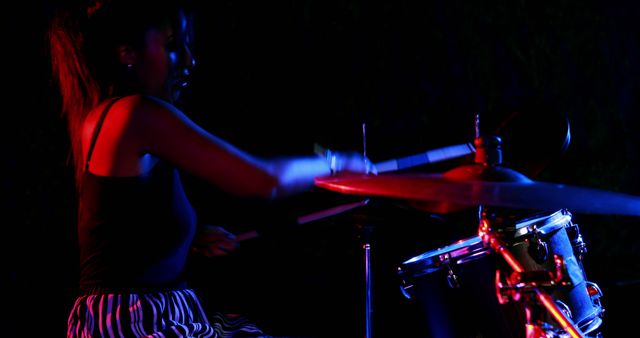 An African American female musician is playing the drums energetically under moody stage lighting, with copy space. Her dynamic performance is captured in a low-light setting, emphasizing the vibrant atmosphere of live music events.
