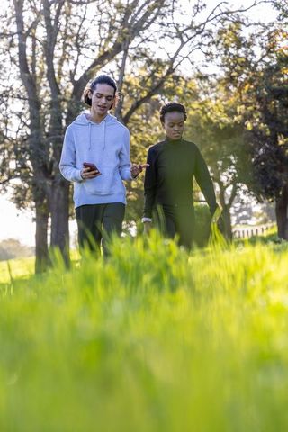 Diverse young adults walking in sunlit park holding smartphone and sharing conversation
