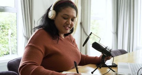 Mid-adult woman recording podcast at home desk with headphones, microphone, laptop, note-taking