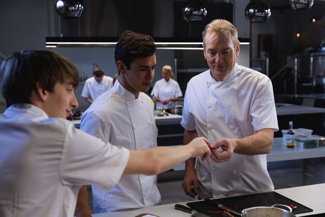 Chefs collaborating in a modern restaurant kitchen, with a student passing ingredients to a male chef. Ideal for use in articles about culinary education, teamwork in professional kitchens, and restaurant training programs.