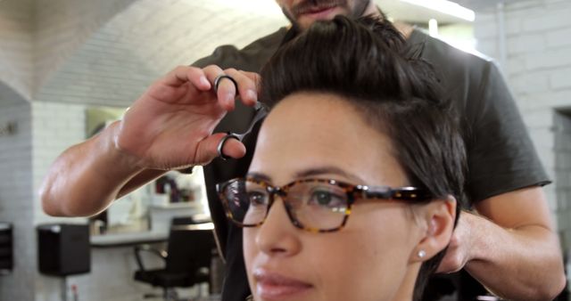 A young Caucasian woman is getting a haircut from a hairstylist, with copy space. She wears glasses and appears relaxed as the professional works on styling her hair.
