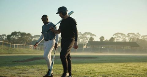 Two baseball teammates walking on misty sunrise outfield carrying bats, one wearing helmet
