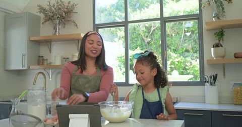 Mother and daughter baking together at kitchen island stirring batter and following tablet