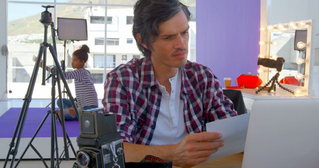 Caucasian man reviews notes in a photography studio. Teenage African American girl prepares in the background, capturing a behind-the-scenes moment.