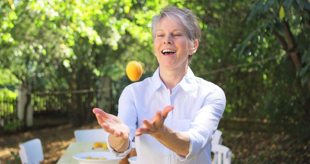 A senior woman joyfully playing with fresh fruit in a garden setting under sunlight, expressing enjoyment and an active lifestyle. Ideal for use in advertisements promoting healthy living, senior well-being, outdoor activities, and fruit-related products.