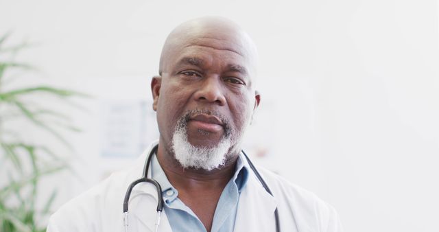 This image depicts a confident African American senior doctor wearing a white coat and stethoscope. He is looking directly at the camera with a serious and professional expression. The background is light and medical-themed, indicative of a hospital or clinic setting. Ideal for use in health-related advertisements, medical websites, educational materials, and healthcare promotions.