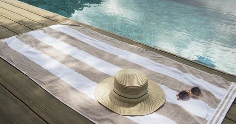 Striped beige towel lying on wooden deck with straw hat and sunglasses by pool
