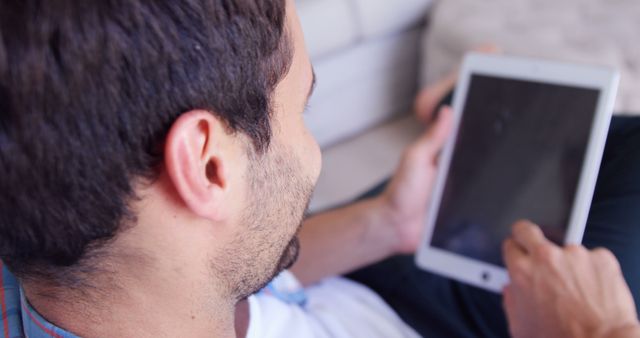 Young, casually dressed man sits at home using a digital tablet. Ideal for illustrating modern technology use or relaxed home environments. Suitable for technology, lifestyle, or remote work themes.