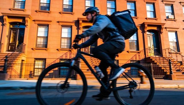 Bike courier riding past brownstone row delivering order during golden hour city commute