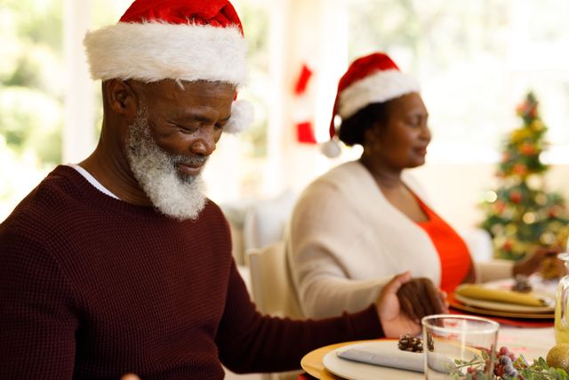 African American couple holding hands and praying at a Christmas dinner table, both wearing Santa hats. Ideal for holiday greeting cards, festive advertisements, family celebration promotions, and articles about holiday traditions and togetherness.