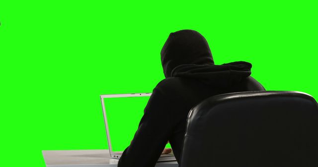 A person in a hooded top is working on a laptop against a green screen background, with copy space. The setup suggests a concept related to technology, privacy, or cybersecurity.