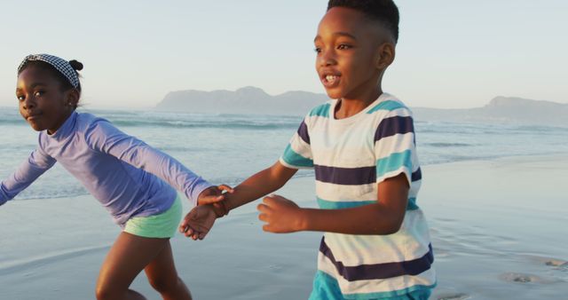 Happy african american siblings holding hands and running on sunny beach. healthy and active time beach holiday.