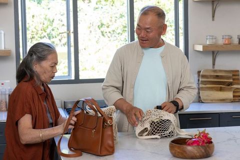 Senior asian couple packing groceries at kitchen island with reusable net bags