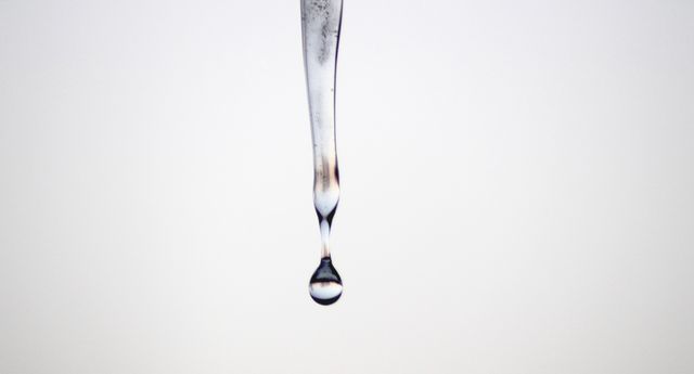 Image shows a single water droplet hanging from the end of a pipette against a white background. It captures the precision and purity of liquid in a scientific context. Ideal for use in educational, scientific, medical, or environmental media to illustrate concepts of precision, purity, and simplicity.