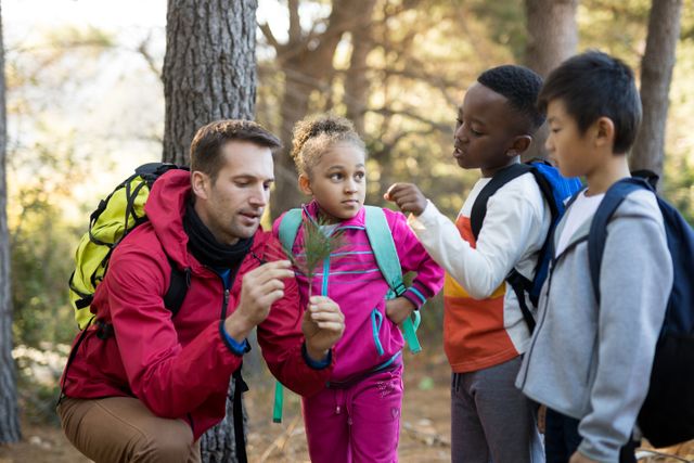 Teacher and a group of diverse children examining a plant in a park. They are surrounded by trees, and all are wearing backpacks, indicating they are on a hike or nature exploration. This image can be used for educational content, promoting outdoor activities, diversity in education, and environmental awareness.