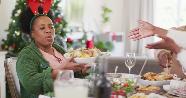 Family gathered around dinner table sharing a roast chicken during Christmas celebration. A woman wearing Christmas antlers passing the roast chicken platter. Background includes a decorated Christmas tree and festive decorations. Useful for holiday marketing, family-oriented campaigns, festive greeting cards, seasonal promotions, and articles on family traditions.