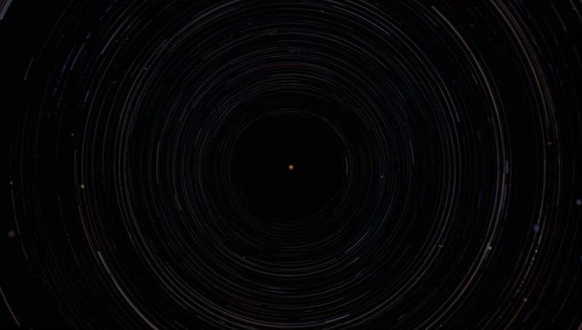 Concentric star trails circling dark celestial core with single orange star point