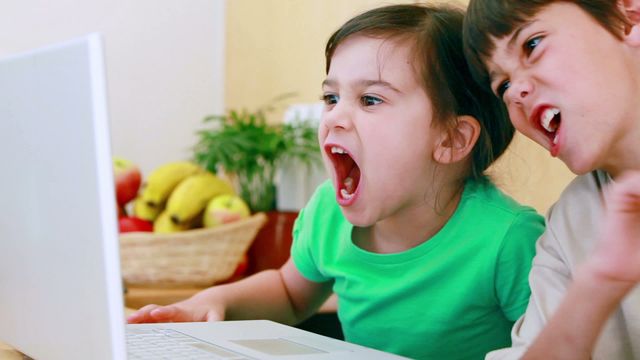 Two children are reacting with excitement and enthusiasm while interacting with their laptop, appearing engaged and joyful. It looks like they are watching something thrilling or enjoying a fun game. This scenario is set in a kitchen environment that includes a fruit basket, adding a cozy and homey element. Suitable for use in themes such as technology interaction among kids, family bonding moments, or depicting fun and educational online activities for children.