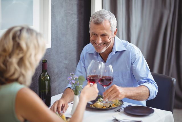 Mature couple enjoying a romantic dinner in a restaurant, toasting their glasses of red wine. Ideal for use in advertisements for restaurants, wine brands, or romantic getaways. Can also be used in articles about relationships, senior lifestyle, and special occasions.